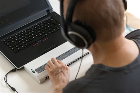A person sitting in front of a laptop and braille display. The person is wearing headphones connected to the laptop and is using their hands to touch and read the braille display.